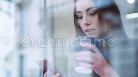 漂亮的年轻女孩穿着时髦的打扮坐在咖啡馆的窗户旁享受美味的热咖啡上网发短信视频的预览图