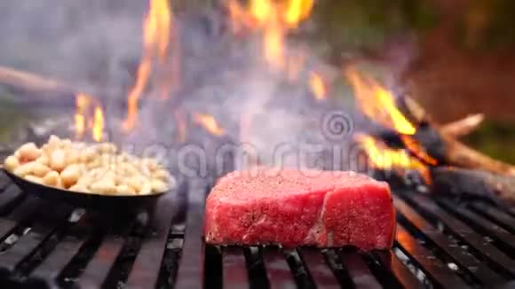 人手把新鲜切好的蔬菜放在黑色烤盘上在森林营地的火焰中放着一大块牛肉牛排粉红色的肉视频的预览图