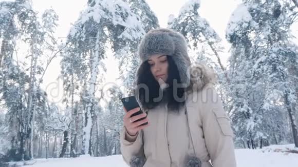戴着温暖帽子的年轻白种人女人在冬天的雪林里用智能手机视频的预览图