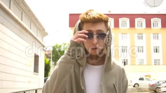 一个穿着时髦衣服的年轻红发男子在城里散步摘下太阳镜的肖像视频的预览图
