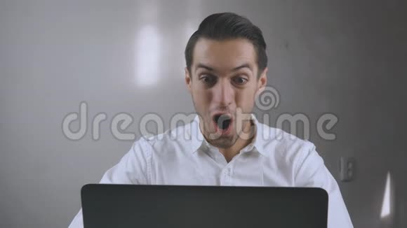 穿着白色衬衫的年轻人非常高兴坐在办公室的笔记本电脑旁他是个胜利者或什么他赢了的东西好的视频的预览图