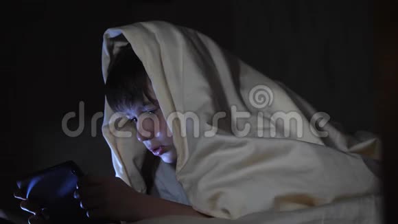 一个孩子晚上在毯子下面使用平板电脑这个男孩玩电脑游戏视频的预览图
