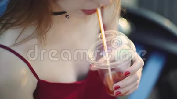 美丽的年轻红发女人坐在公园的长凳上喝果汁在日落时使用智能手机视频的预览图