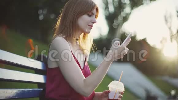 美丽的年轻红发女人坐在公园的长凳上喝果汁在日落时使用智能手机视频的预览图