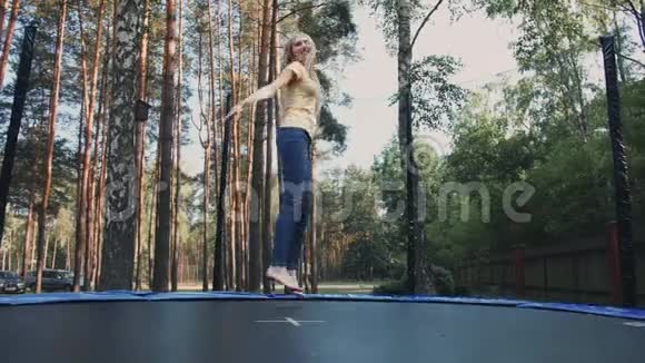 快乐的女性在蹦床上跳跃年轻漂亮的金发赤脚女人穿着浅色的夏季衬衫和牛仔裤视频的预览图