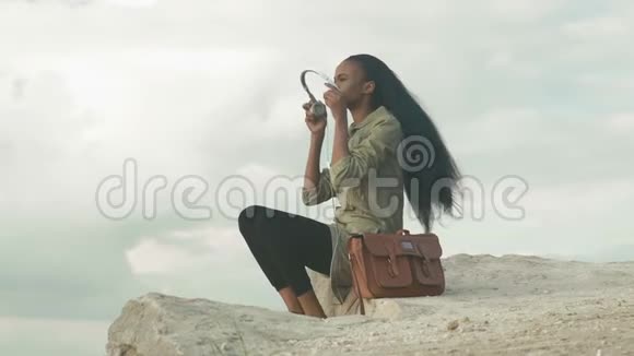 美丽的非洲裔美国年轻女性带着智能手机和耳机在蓝天和云朵上听音乐视频的预览图