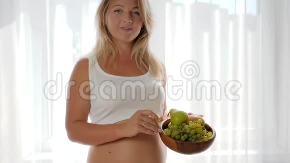 怀孕肚子大的女性在窗户附近吃葡萄拿着木盘视频的预览图