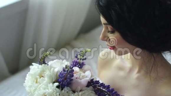 一个温柔迷人的黑发女人的早晨美丽的新娘微笑着看着窗外和一束花视频的预览图