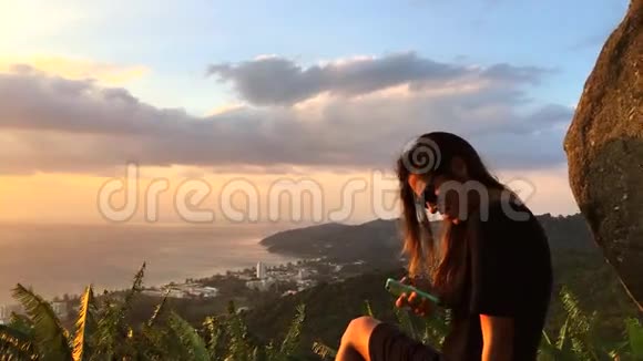 美丽的女人坐在悬崖上在智能手机上发短信泰国普吉岛高清慢动作惊人的视野视频的预览图