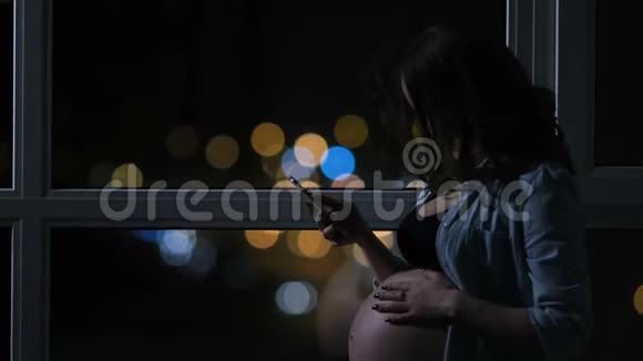 孕妇在晚上用手机说话摸着他的肚子站在大的全景窗口视频的预览图