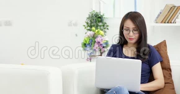戴眼镜的年轻女性坐在沙发上在家里使用笔记本电脑在生活背景下工作室内视频的预览图