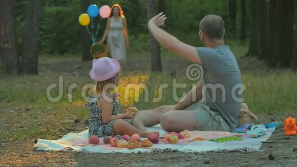 爸爸和小女孩在公园的毯子上放松等待带篮子的女人视频的预览图