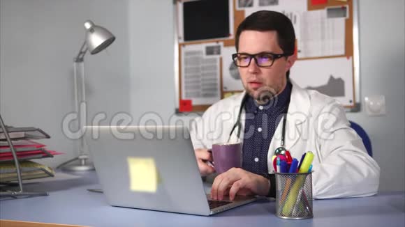 穿着白大褂的医生正在工作场所喝茶看着笔记本电脑视频的预览图