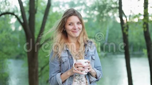 年轻害羞的长发女孩手里拿着一杯茶或咖啡在模糊的自然背景下漂亮女人视频的预览图