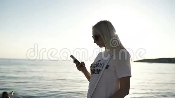日落时分女孩在海洋海岸的智能手机上打字视频的预览图