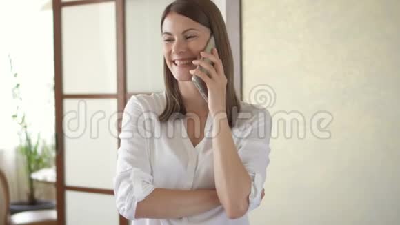 穿着白色上衣的休闲年轻女子站在客厅里笑着笑着打电话视频的预览图