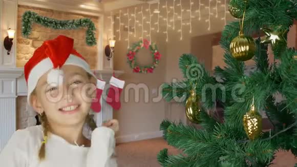 圣诞老人戴着红帽子在圣诞树旁跳舞的快乐少女快乐的白种人女孩在室内近距离按比例分配视频的预览图