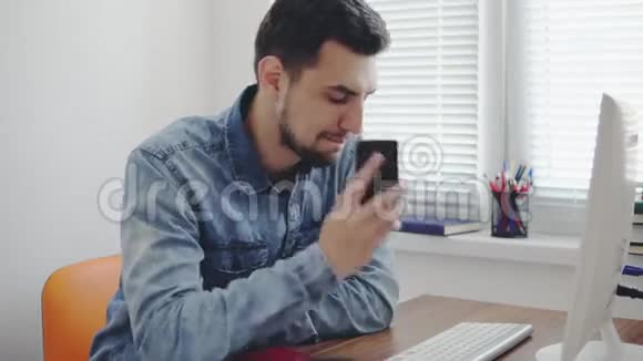 年轻的办公室工作人员拨了一个电话号码用他的手机在办公室里用电脑坐在桌子旁交谈视频的预览图