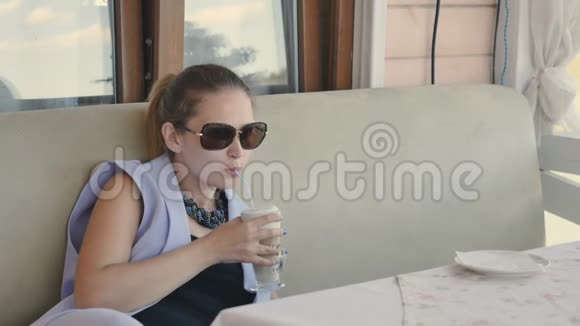 戴太阳镜的漂亮女孩在夏季露台的咖啡厅喝拿铁视频的预览图