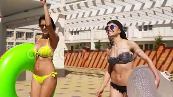 两个戴着太阳镜的迷人女孩在游泳池旁走着充气管微笑着有游泳池的漂亮女人视频的预览图