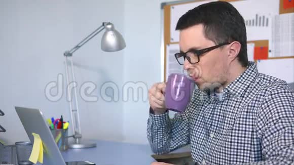 一个男人正在工作场所后面的杯子里喝茶看着笔记本电脑视频的预览图