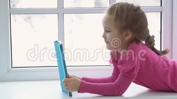 穿着粉红色连衣裙的小女孩正拿着一块平板电脑坐在窗边侧视图视频的预览图