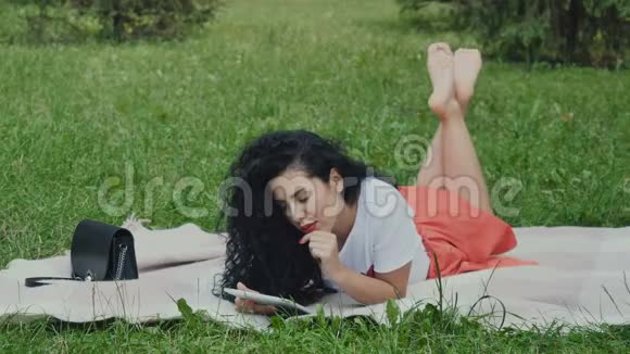 可爱的卷发女孩在公园的格子布上用平板电脑放松视频的预览图