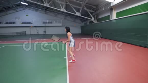 穿着运动服装在室内球场打网球的苗条女子的全长肖像配有专业设备视频的预览图