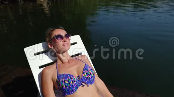 女人戴着太阳镜和泳衣躺在日光浴床上女孩在洪水木水下码头休息特写自我画像视频的预览图