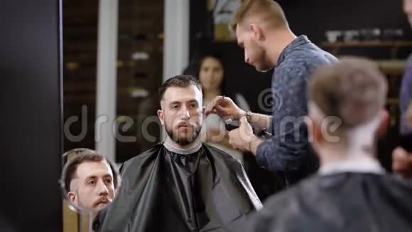 时尚的理发师正在刮太阳穴理发店里坐在扶手椅上看着一个留胡子的人的理发器视频的预览图