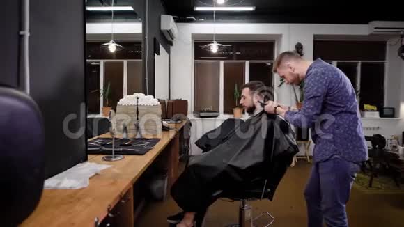 年轻的理发师在扶手椅上走来走去在理发店里和剪刀一起理发有胡子的男人坐在视频的预览图