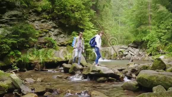 一对带背包的年轻游客穿过森林中的一条山河视频的预览图