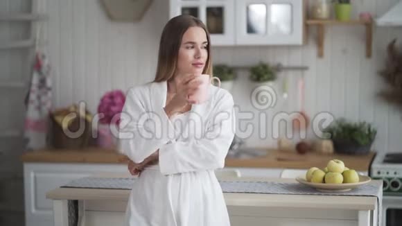 穿着白色浴袍在厨房喝咖啡或茶的年轻女人的肖像视频的预览图