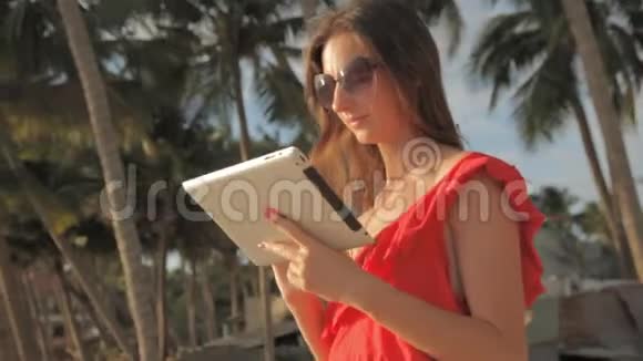 穿着红色连衣裙在阳光明媚的海滩上散步的年轻女子她在用平板电脑微笑着视频的预览图