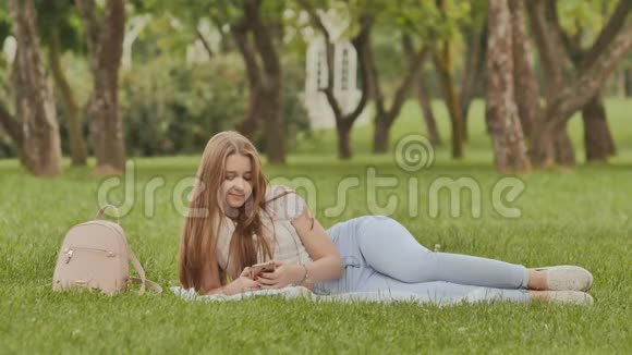 有魅力的年轻女孩带着智能手机躺在草地公园里在学习期间休息视频的预览图