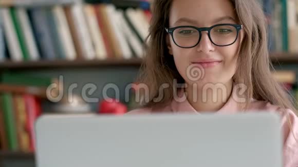 女学生坐在书架旁在图书馆里使用笔记本电脑视频的预览图