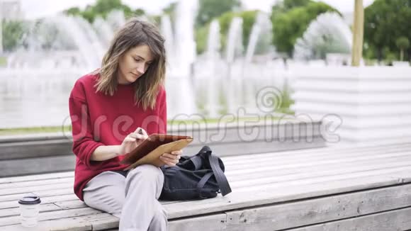 穿红衣服的年轻女子在喷泉旁用平板电脑工作视频的预览图
