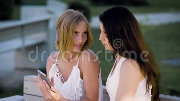 两个年轻时尚的女人坐在长凳上在夏天看智能手机视频的预览图