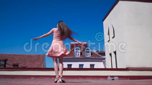 一个穿着粉红色裙子的女孩在院子里旋转在背景蓝天和屋顶的房子慢镜头画面视频的预览图