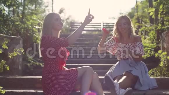 两个迷人的年轻金发女郎在公园里吹泡泡日落时很开心女朋友在公园的台阶上视频的预览图