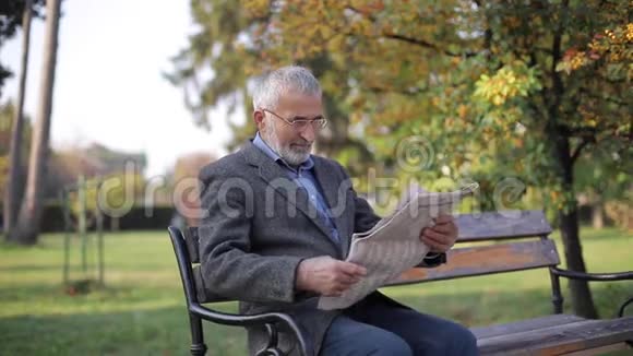 大胡子老人戴眼镜在秋天公园看报纸英俊的白发男子早早地坐在长凳上视频的预览图