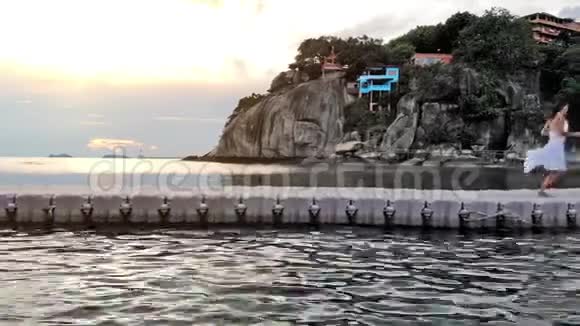 在美丽的日落中年轻的黑发女子穿着白色连衣裙在热带岛屿的海上大桥上奔跑视频的预览图