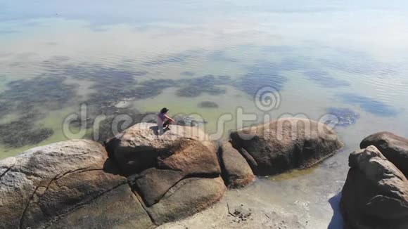 戴帽子的年轻人在海边享受自由独自坐在岩石上从无人机飞到大海的视野1920x1080视频的预览图