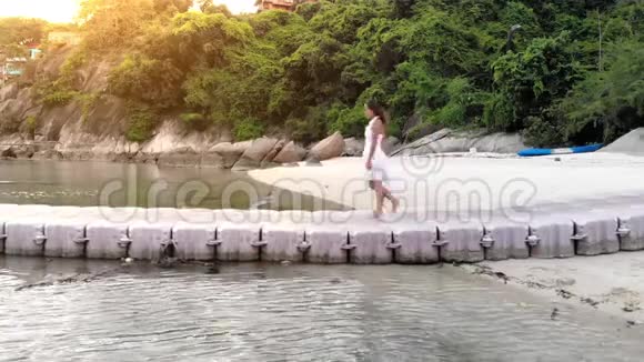 在美丽的日落中年轻快乐的漂亮女人穿着白色连衣裙在热带岛屿的海中桥上奔跑视频的预览图