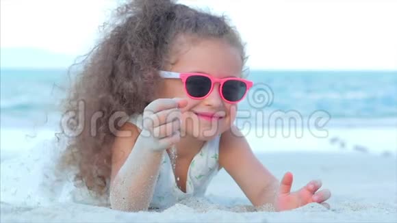 一个戴着粉红色眼镜的漂亮小女孩的特写肖像可爱的微笑着看着相机躺在沙滩上视频的预览图