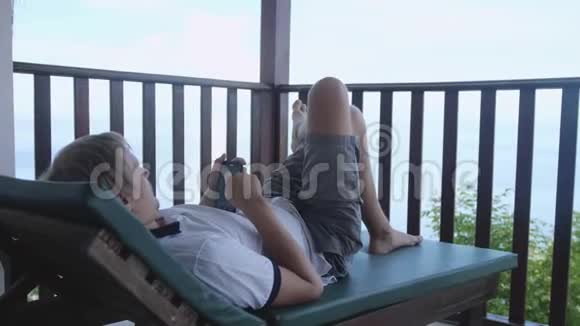 年轻人躺在露台上一边玩电话一边俯瞰大海慢动作3840x2160视频的预览图