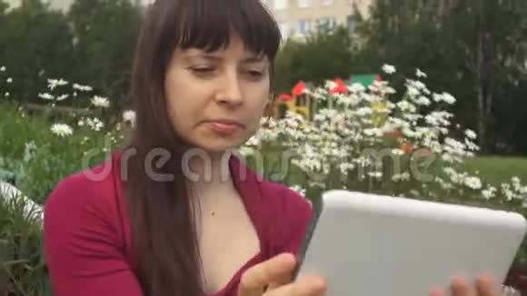 年轻女子使用白色平板电脑坐在户外雏菊旁边视频的预览图