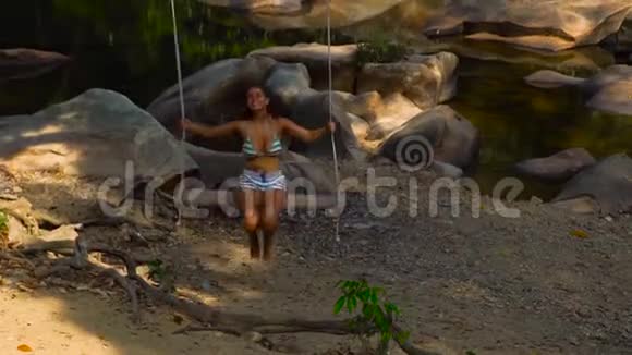 年轻女子在石河岸边摇摆快乐的女人在背景丛林景观的河岸上荡秋千视频的预览图