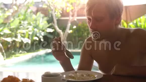 年轻的帅哥吃着燕麦粥喝着茶早餐坐在他的别墅里在游泳池的背景上视频的预览图