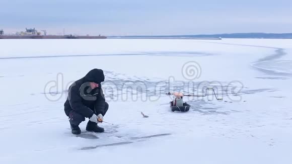 穿着深色衣服的老渔夫在冰冻的河上钓着冬天的鱼竿视频的预览图